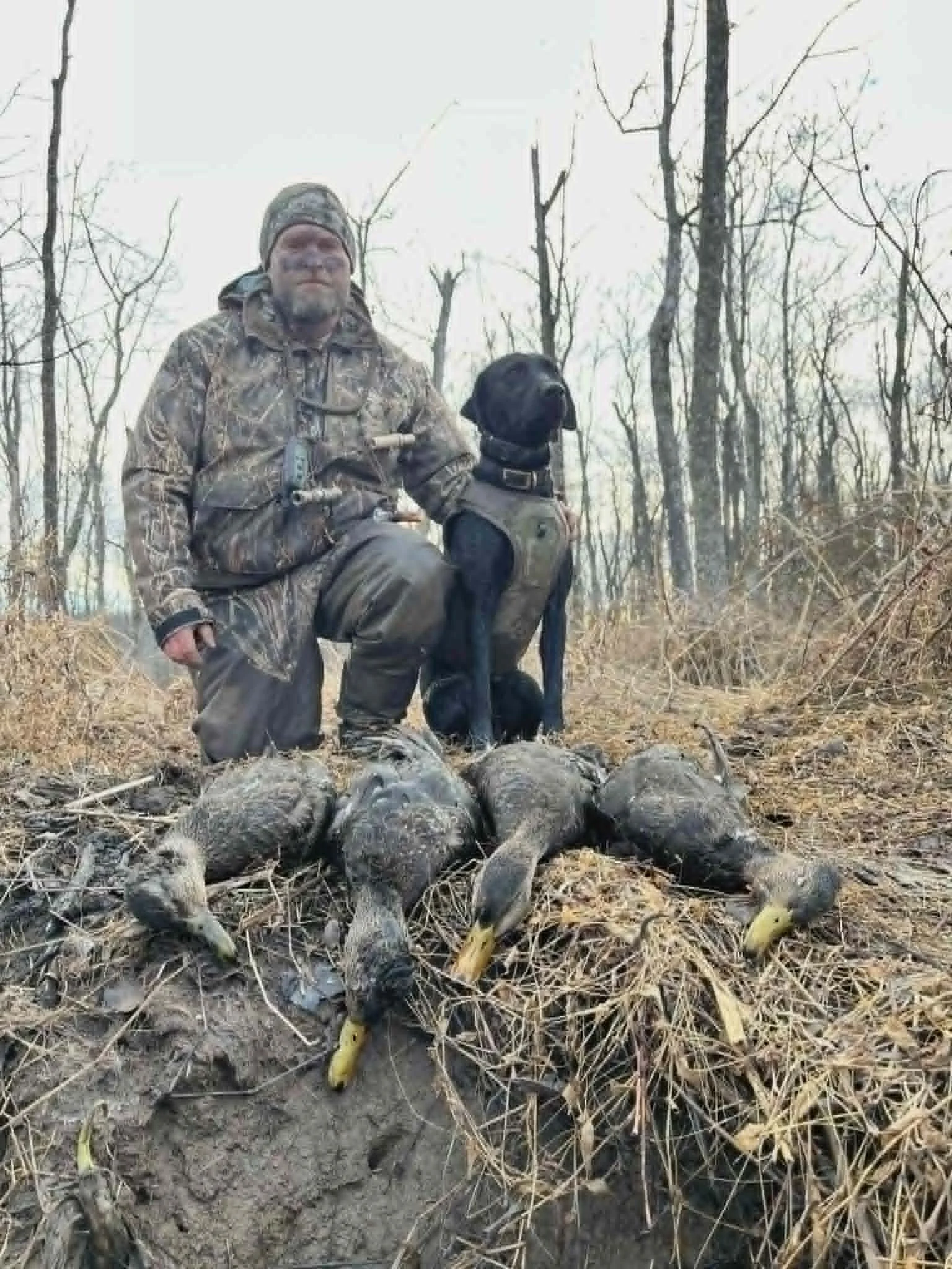 A man in camouflage outdoor clothing kneels in a wooded area with a black dog beside him. In front of them, there are several dead animals on the ground, possibly wild game. The scene appears to be in late fall or winter, with bare trees and dry grass.