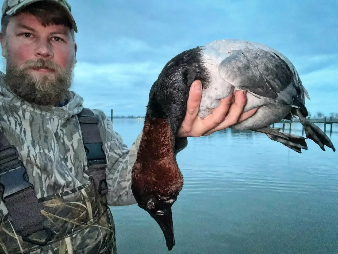 A man with a beard wearing camouflage clothing and gear, holding a large wooden-duck decoy in front of a body of water during dusk.
