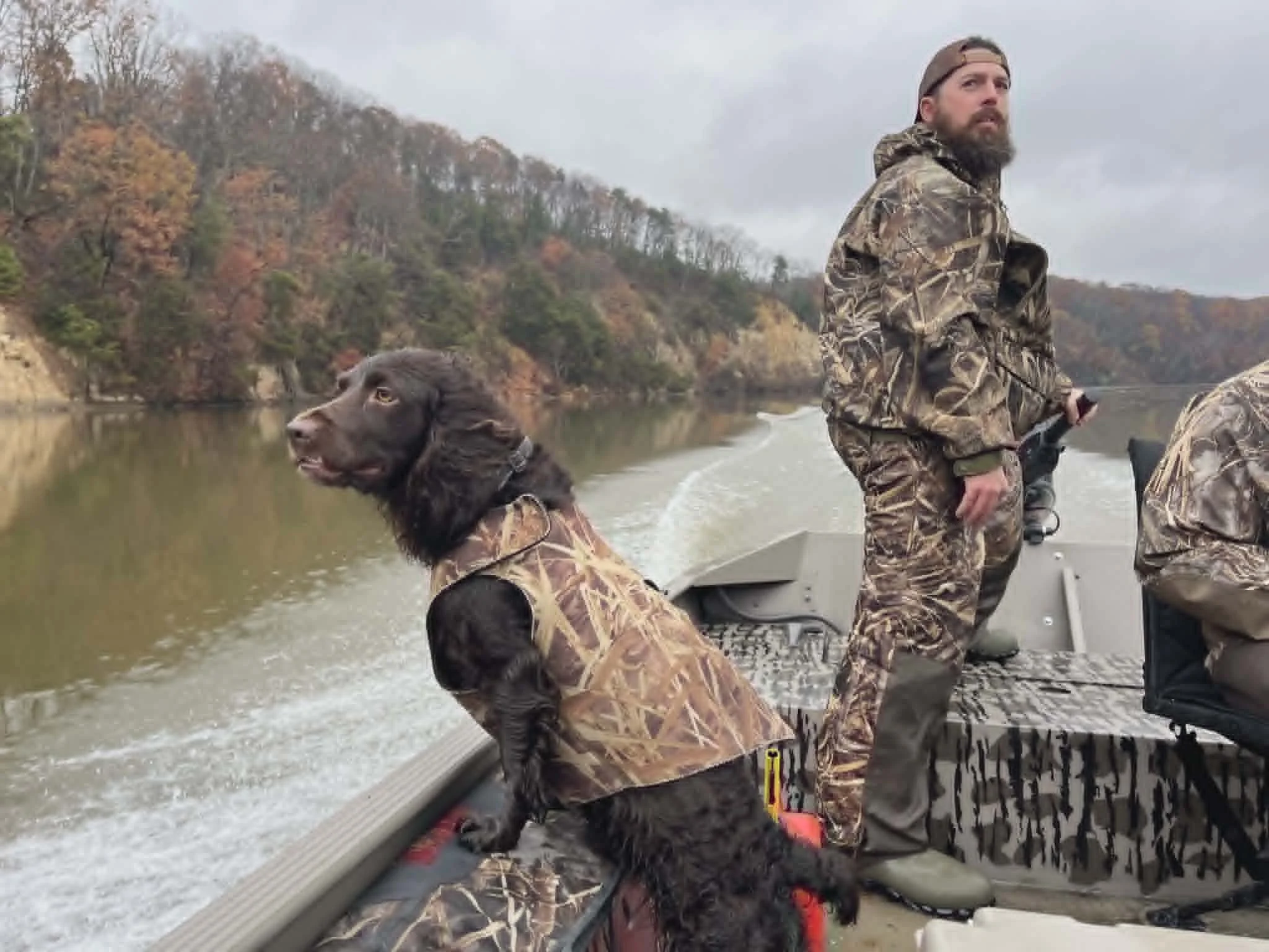 A man with a beard dressed in camouflage clothing and a baseball cap is fishing from a boat on a river, accompanied by a large dog also dressed in camouflage. The river is surrounded by trees with fall foliage and the sky is overcast.