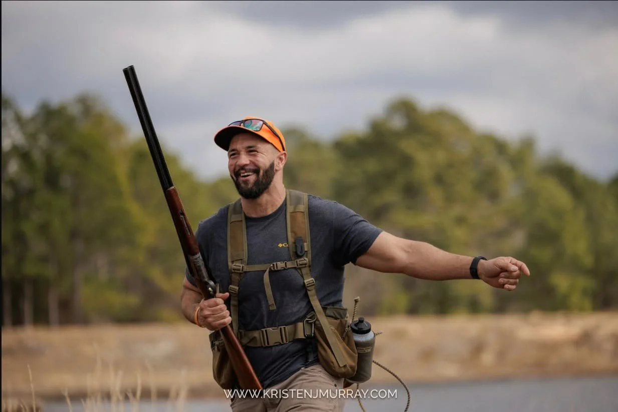 A man is smiling while holding a shotgun outdoors during the daytime, with trees and cloudy sky in the background.