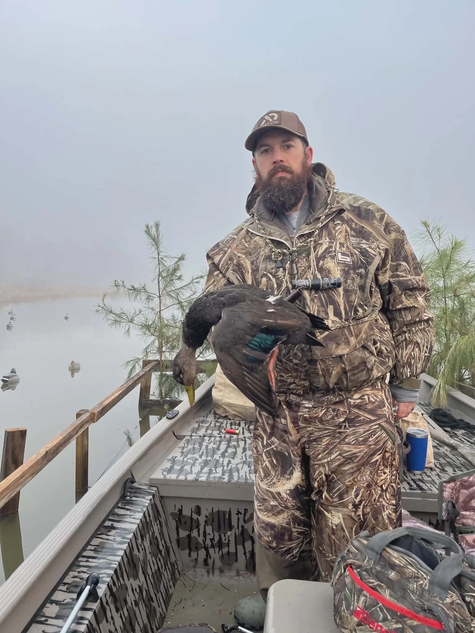 Man in camouflage clothing holding a large bird on a boat, with foggy background and small trees.