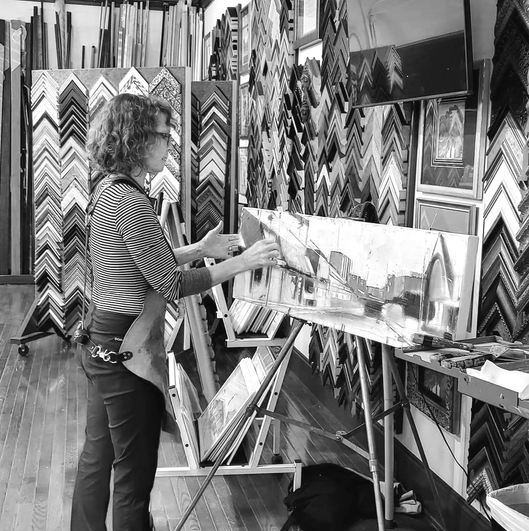 A woman in a striped shirt and glasses is working on a painting in an art store. She is standing in front of an easel with a reflective cityscape painting, surrounded by racks of framed pictures and artwork.