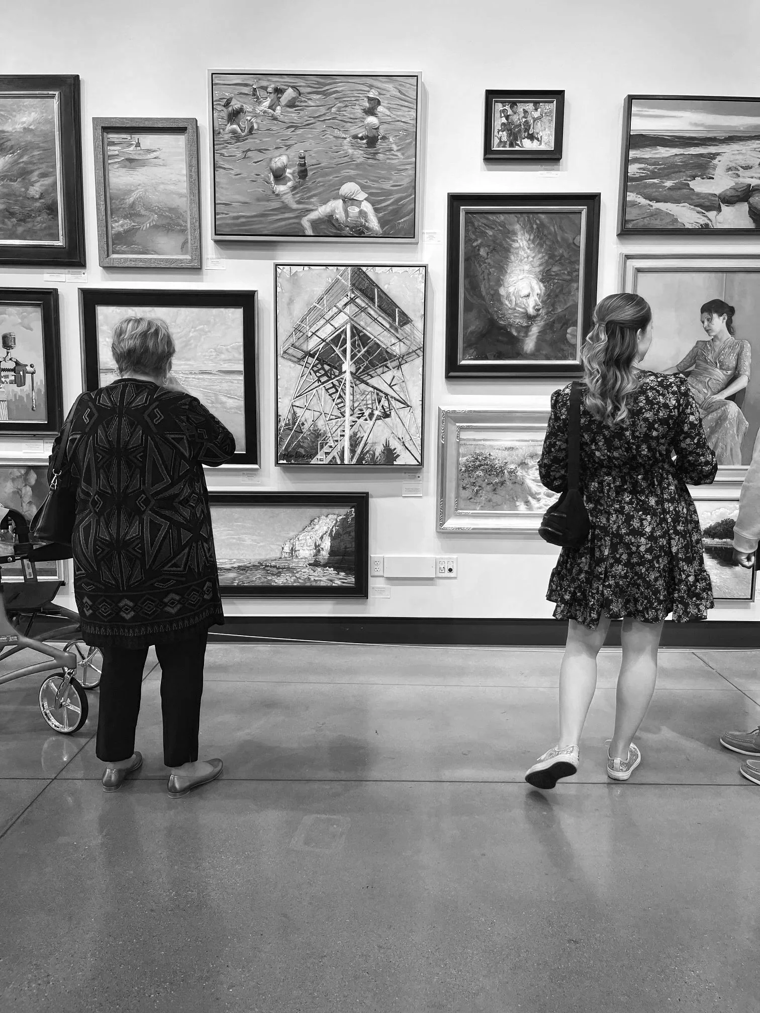 In an art gallery, two women are viewing paintings on the wall. One woman, with her back to the camera, has curly hair and is wearing a black floral dress with white shoes, carrying a black purse. The other woman, with short hair, is wearing a patterned coat and standing near a roller walker, looking at a painting of a seascape. The wall displays various paintings, including a large one of people swimming and others depicting landscapes.