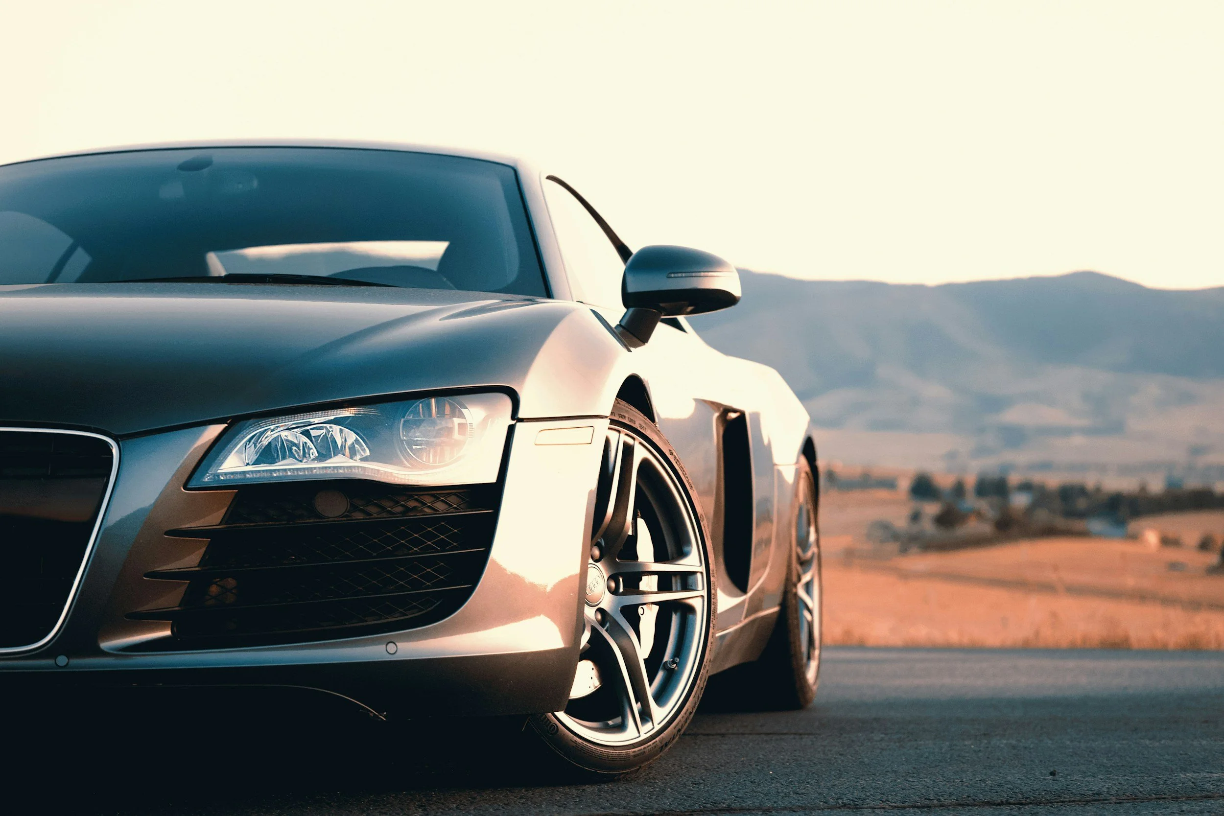 Close-up of the front part of a silver sports car on a road, with a distant desert landscape and mountains under a clear sky in the background.