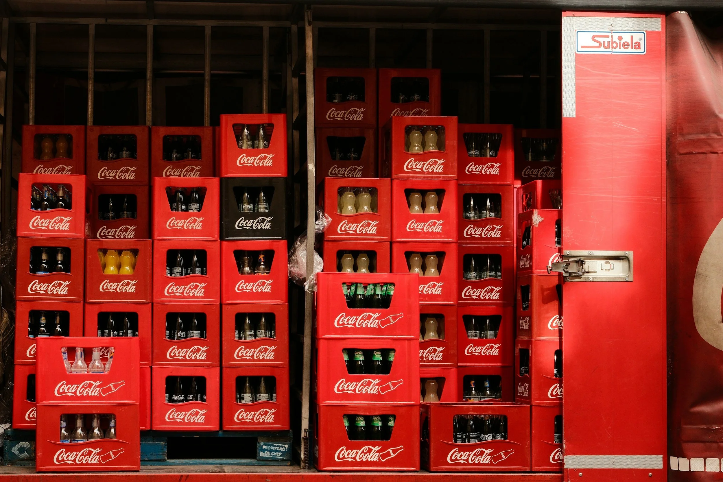 Shelves with multiple red Coca-Cola crates stacked, containing glass bottles and cans of soda, next to a red Subiela vending machine.