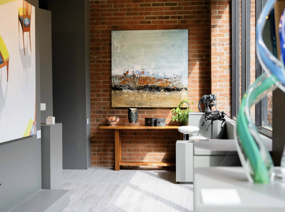 An art gallery corner with a large abstract painting on a brick wall, a wooden table with decorative items, and sculptures near a window with natural light.