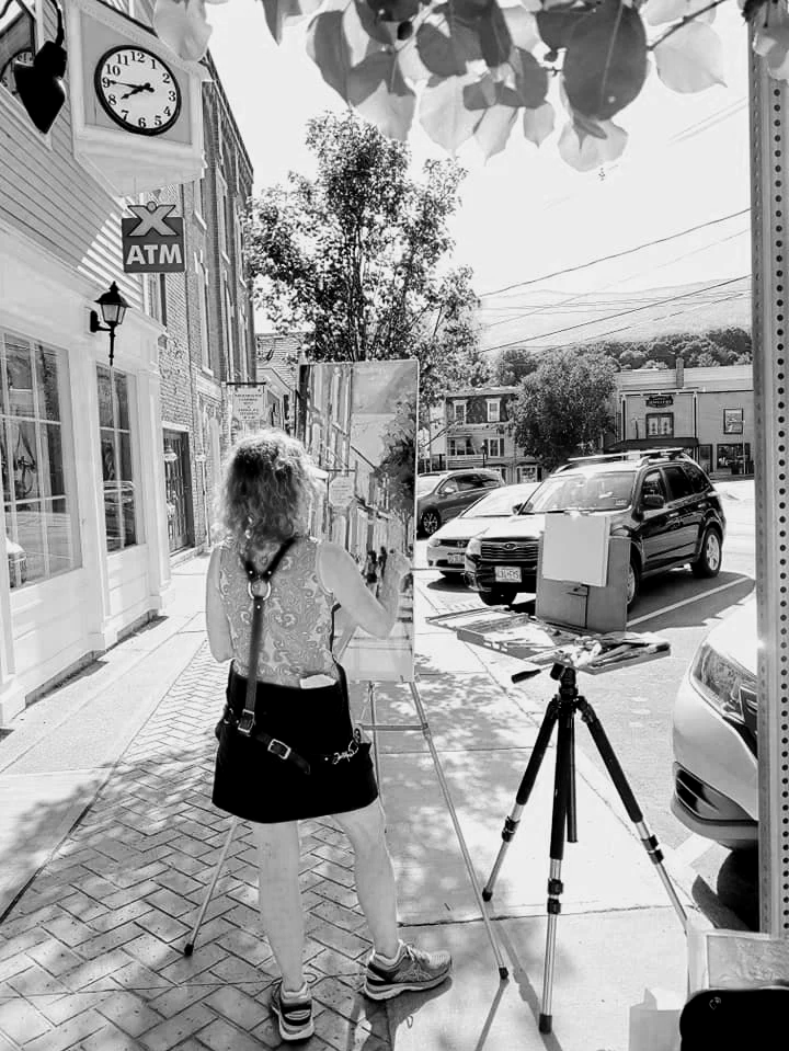 A woman painting outdoors on a city sidewalk with an easel, surrounded by parked cars and buildings, under a tree with dappled sunlight.