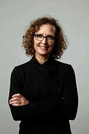 A woman with curly hair, glasses, and a black top smiling, standing with arms crossed against a plain light background.