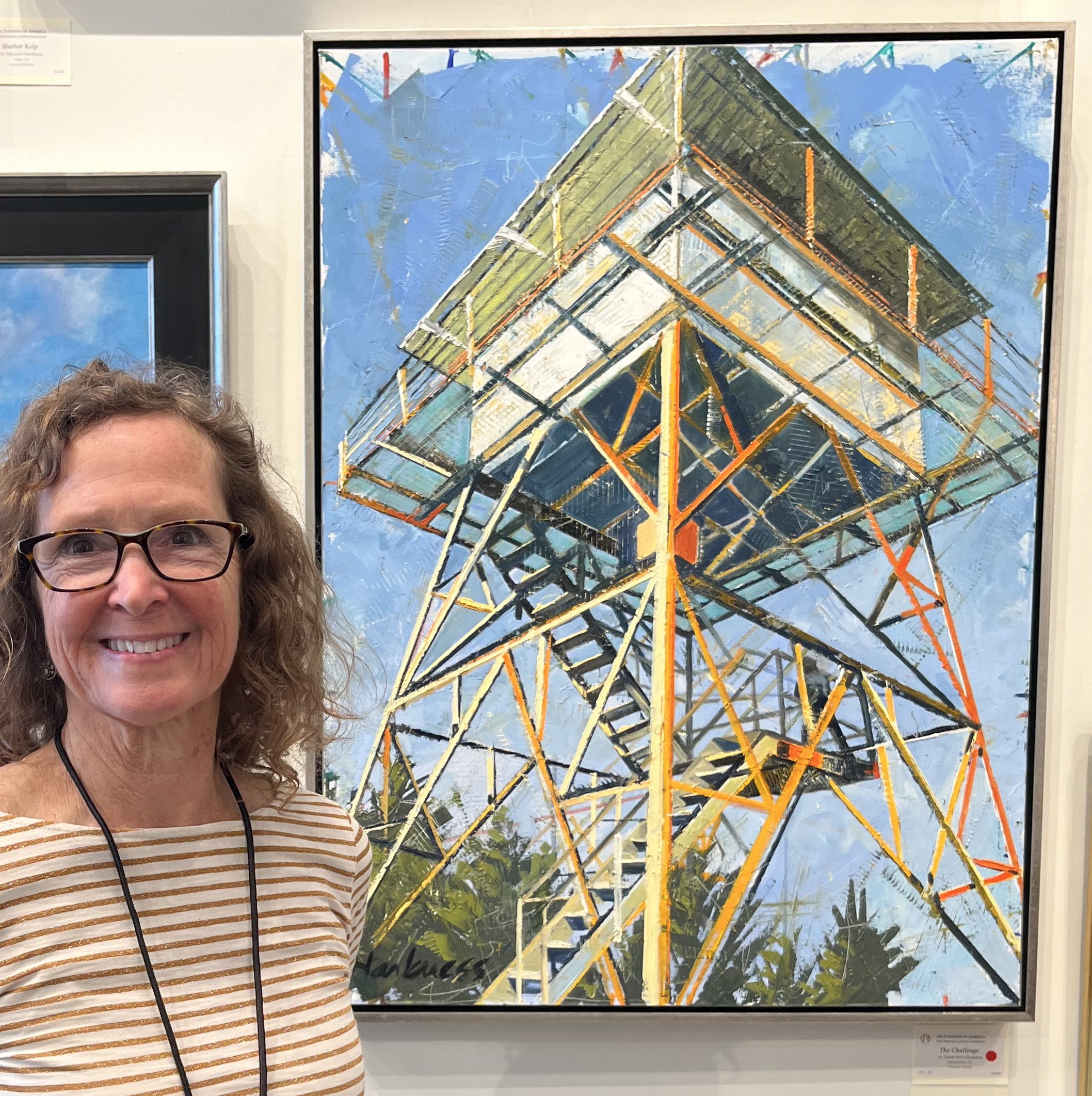 Woman with glasses wearing a striped shirt standing in front of a large framed artwork depicting a colorful geometric architectural structure, displayed at an art gallery.