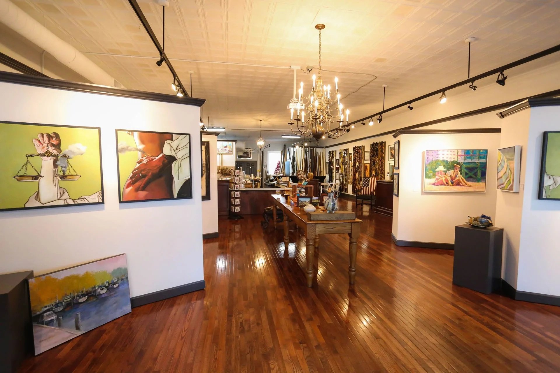Interior of an art gallery with paintings on white walls, wooden flooring, a chandelier, and display tables.