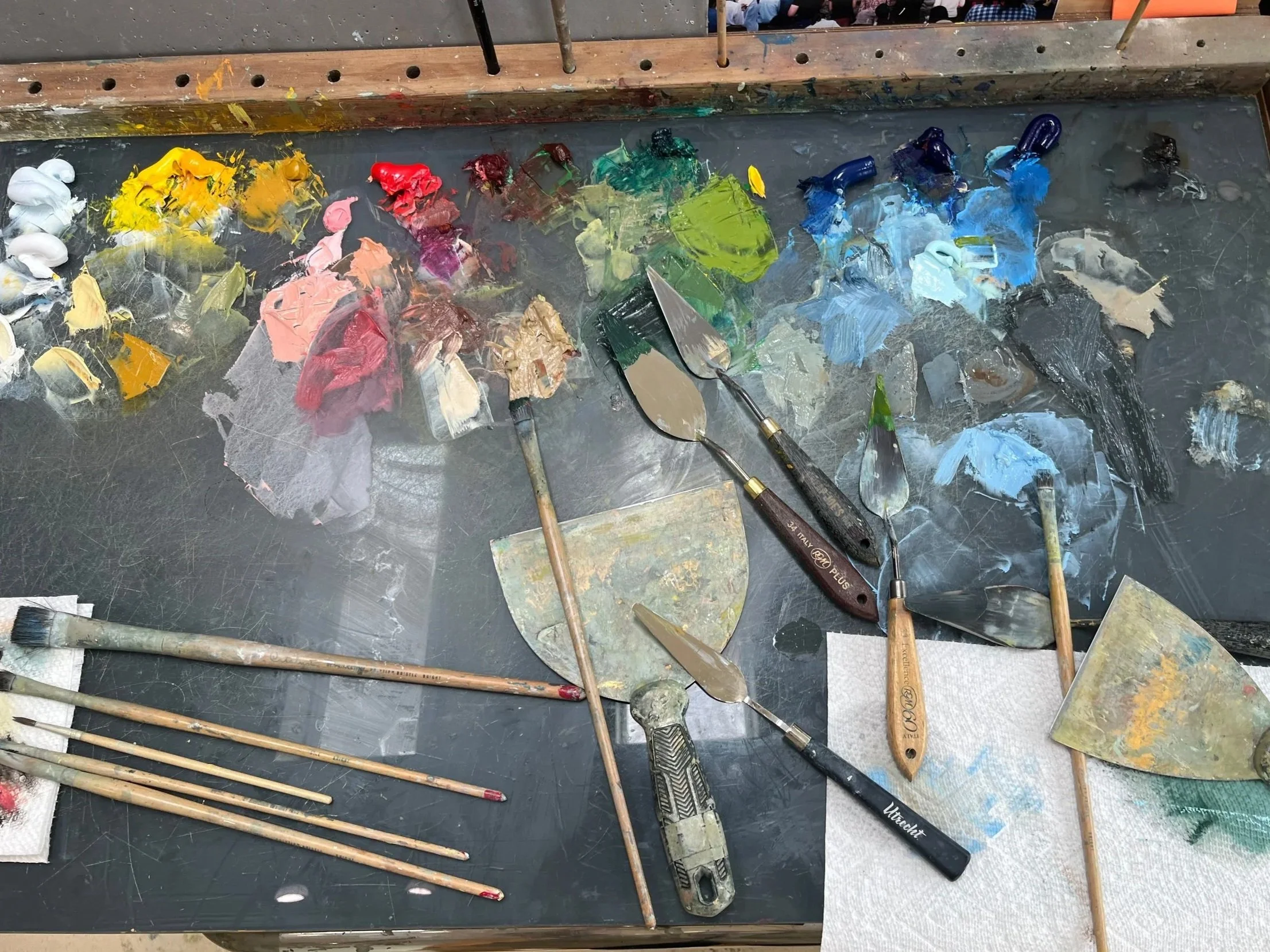 A painter's workspace with various paint smudges, brushes, spatulas, and palette knives scattered across a black work surface, with a background showing a stage or workshop area with people.