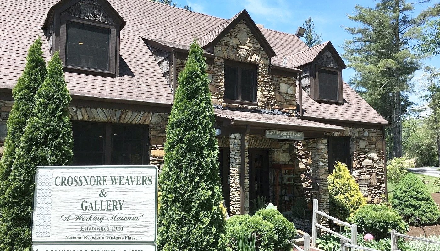 Stone building with a sign that reads "Crossnore Weavers & Gallery" and indicates it is "A Working Museum" established in 1920, surrounded by greenery including bushes and tall trees.