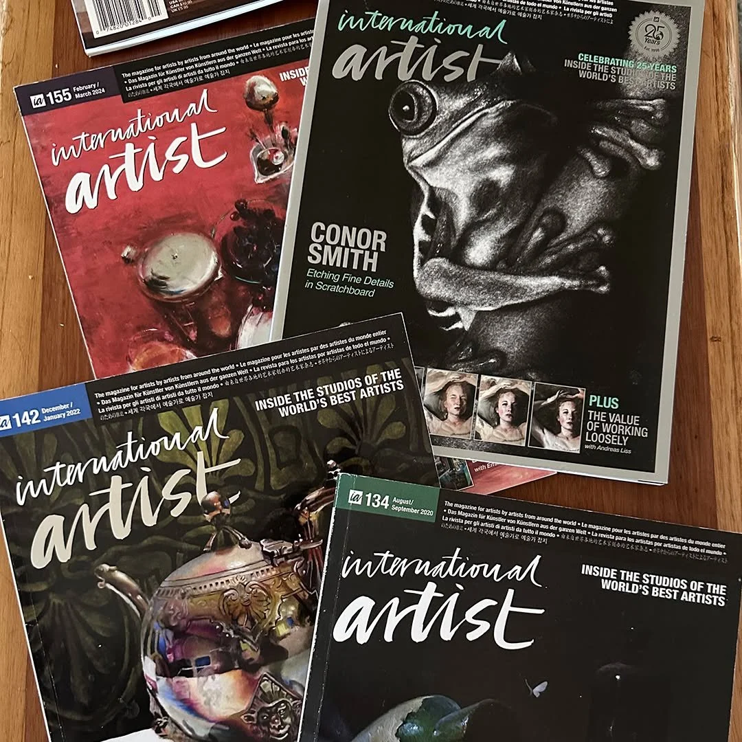 Four issues of International Artist magazine laid out on a wooden surface. The covers feature various artworks including a painted still life, a detailed scratchboard of a frog, a colorful glass teapot, and a dark background with a butterfly and a bird.