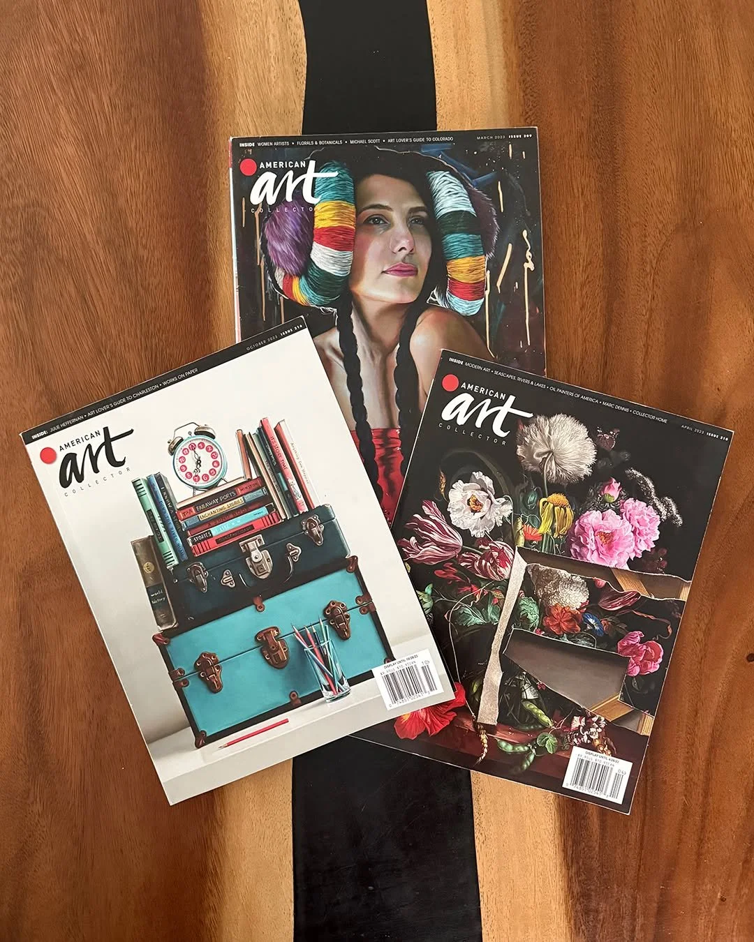 Three issues of American Art magazine with artistic covers, one featuring a woman with colorful hair and braids, another with floral artwork, and the third with a vintage suitcase filled with books and a clock, are spread on a wooden table with a black stripe.