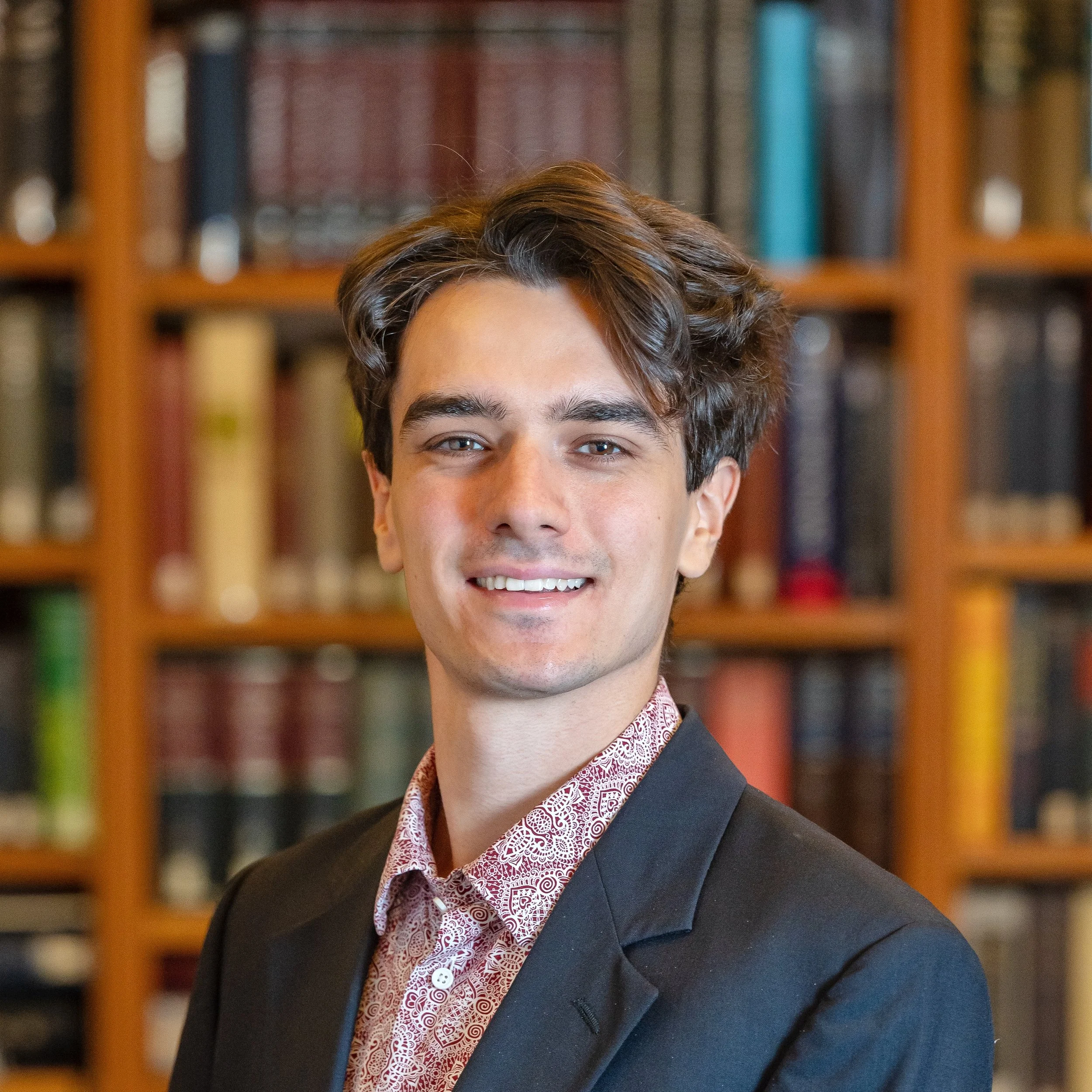 Close-up of young man with dark hair, blue eyes, smiling, wearing a suit and patterned shirt, in front of a bookshelf filled with books.