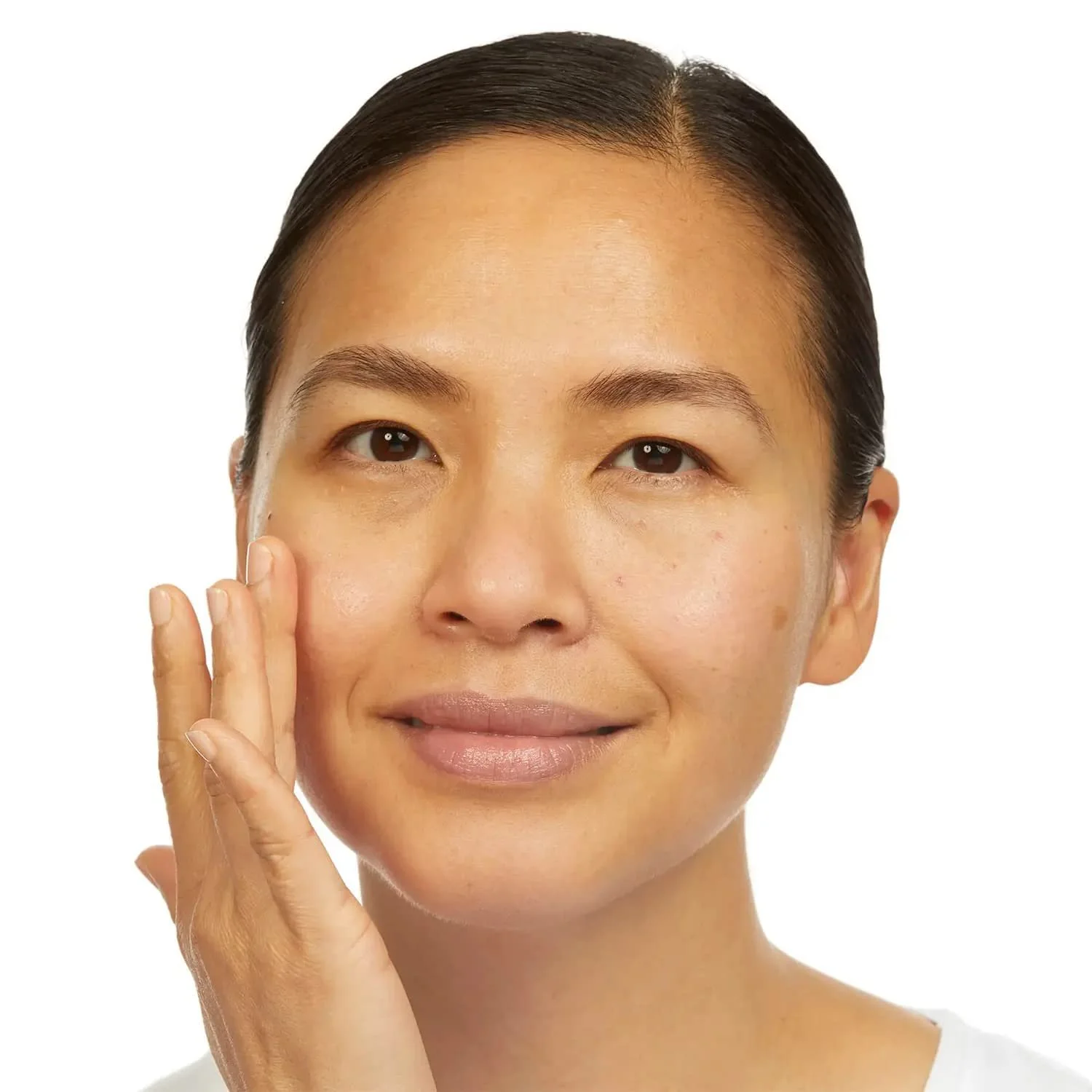 A woman with dark hair, touching her face gently with her fingers, with clear skin and a slight smile, against a white background.