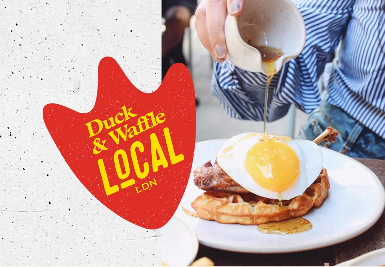 A person pouring syrup over a breakfast dish with waffles, fried chicken, and sunny-side-up eggs at a restaurant.