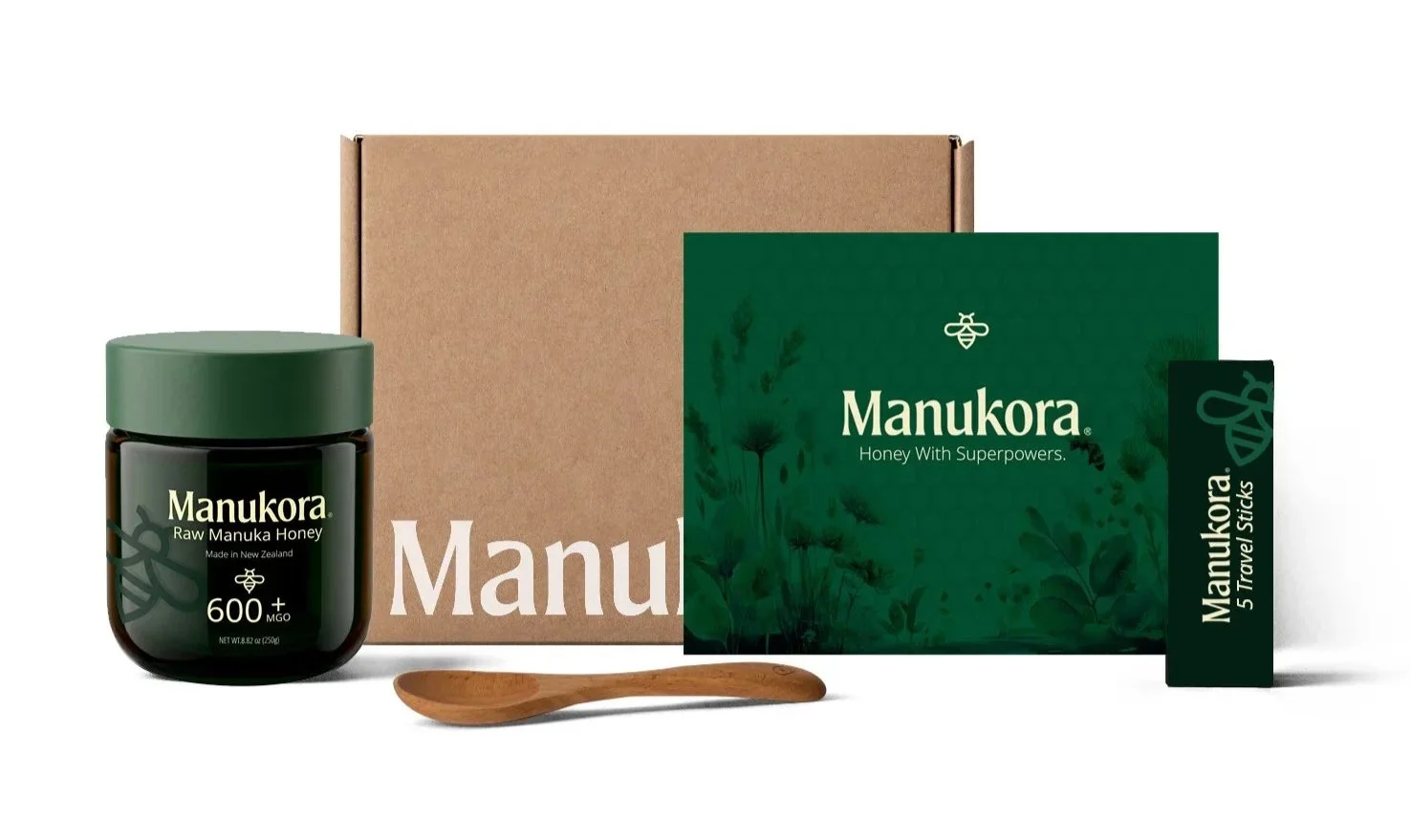 Manukora brand raw manuka honey jar, cardboard box, green travel stick pack, brochure, and wooden spoon on a white background.