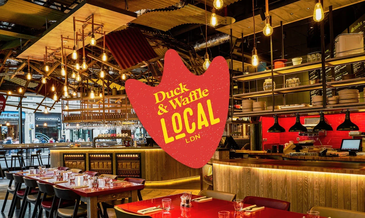 Interior of a cozy restaurant with hanging light bulbs, a bar area with shelves of plates, glasses, and condiments, and a red brick accent wall. Red and black chairs and tables are set for customers, and there's a neon sign outside the window.
