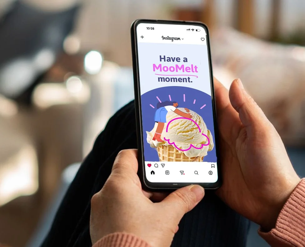 Hand holding a smartphone with an Instagram post showing a cartoon girl with orange hair and blue dress hugging a large scoop of vanilla ice cream in a cone. The post says 'Have a MooMelt moment.'