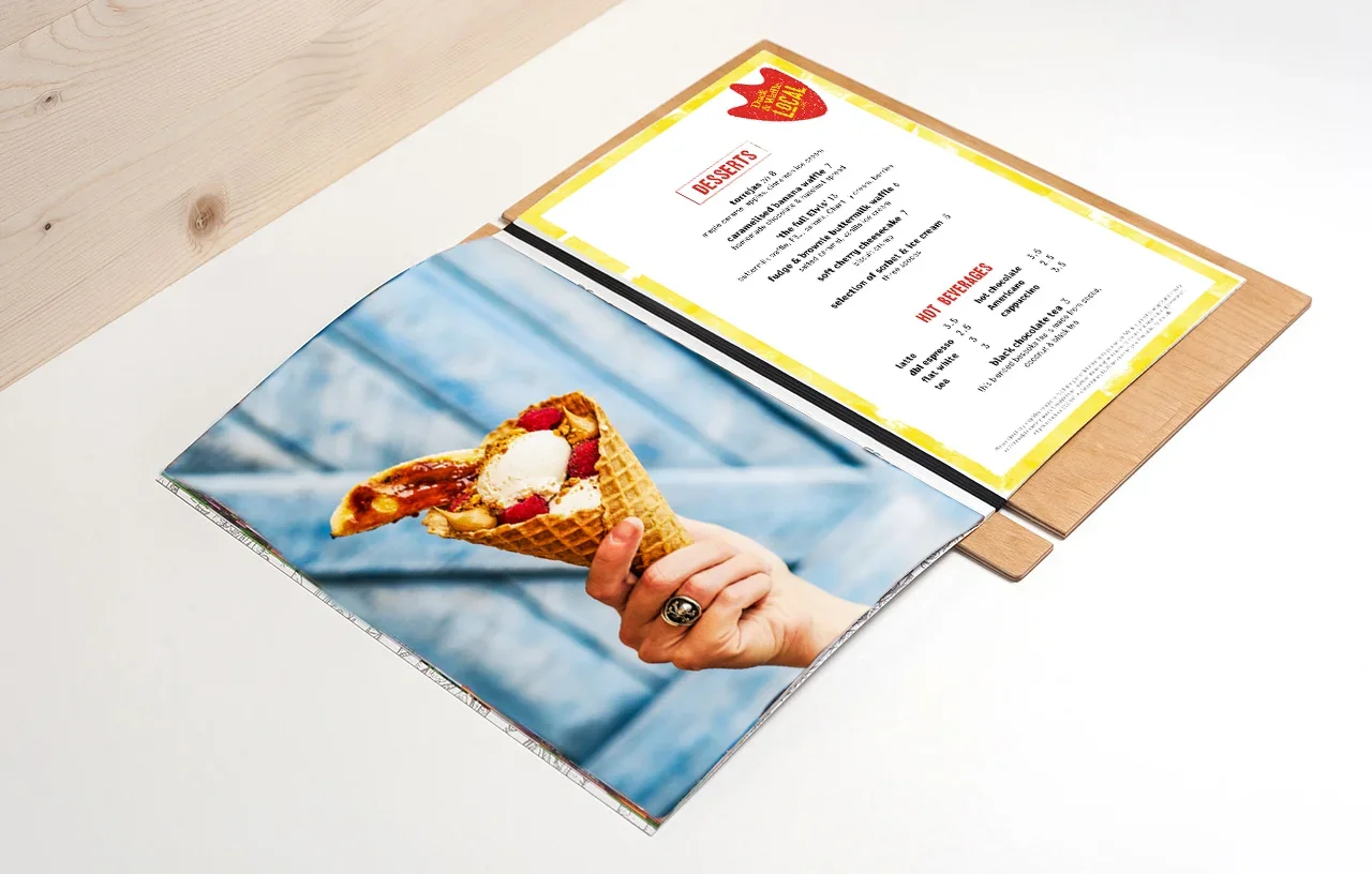 Open magazine with a photo of a person holding a waffle cone with ice cream and strawberries on an outdoor table, with a menu on top of the magazine.