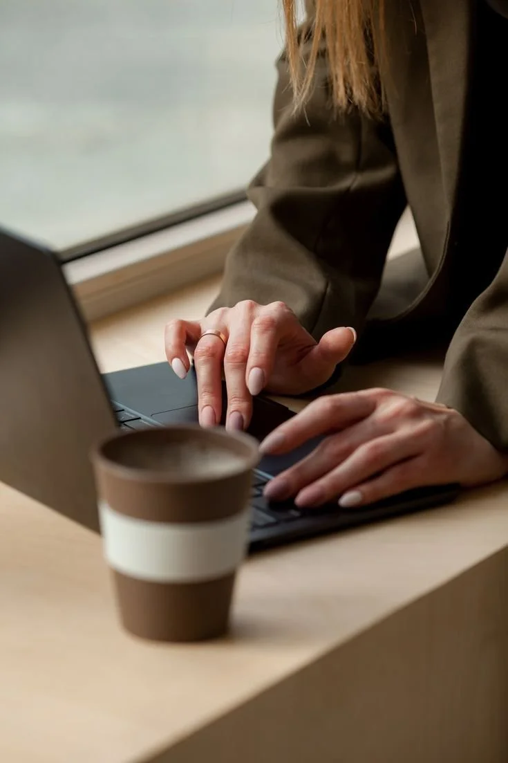 Person working on a laptop with a coffee cup nearby, sitting near a window.
