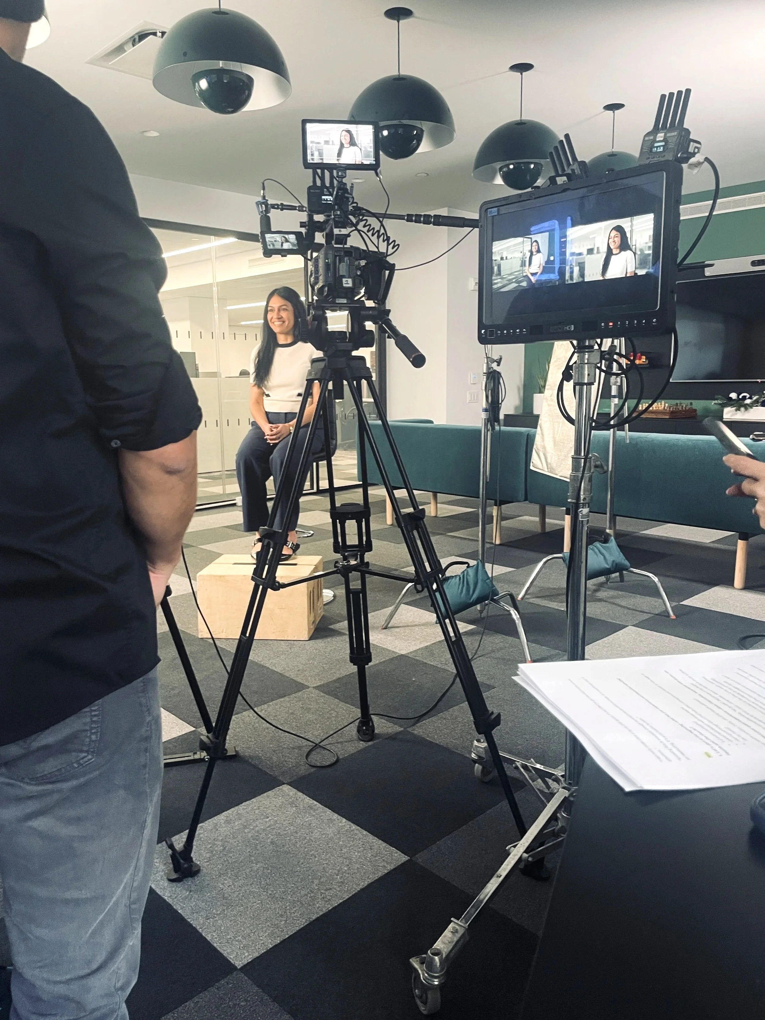 Recording or filming set in an office with a woman smiling and sitting on a stool, surrounded by professional camera equipment, lights, and monitors.