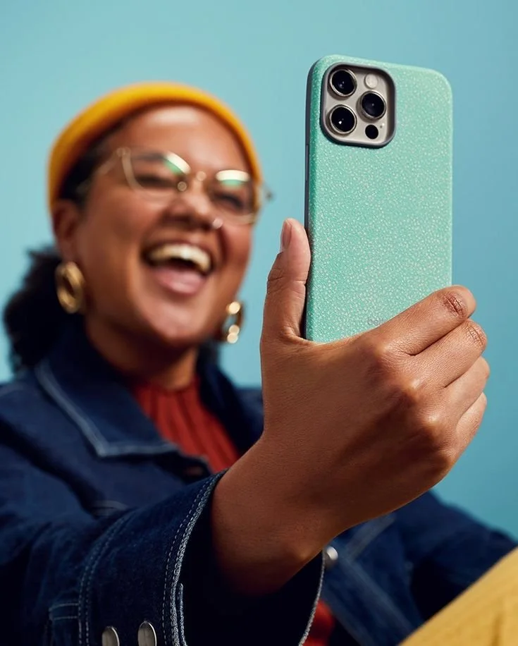 Person taking a selfie with a mint green smartphone, smiling, wearing glasses, a yellow beanie, and a denim jacket against a blue background.