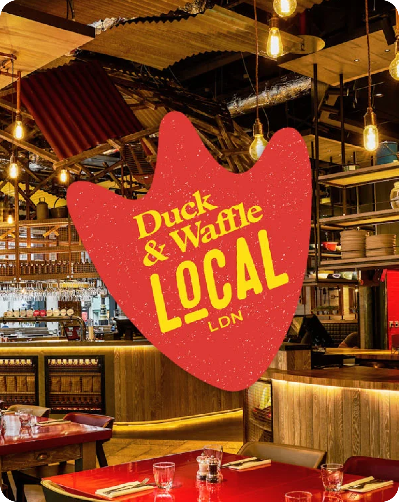 Interior of a restaurant with a large red sign hanging that reads "Duck & Waffle Local LDN." The restaurant features warm lighting, wooden accents, and dining tables set with glasses and utensils.