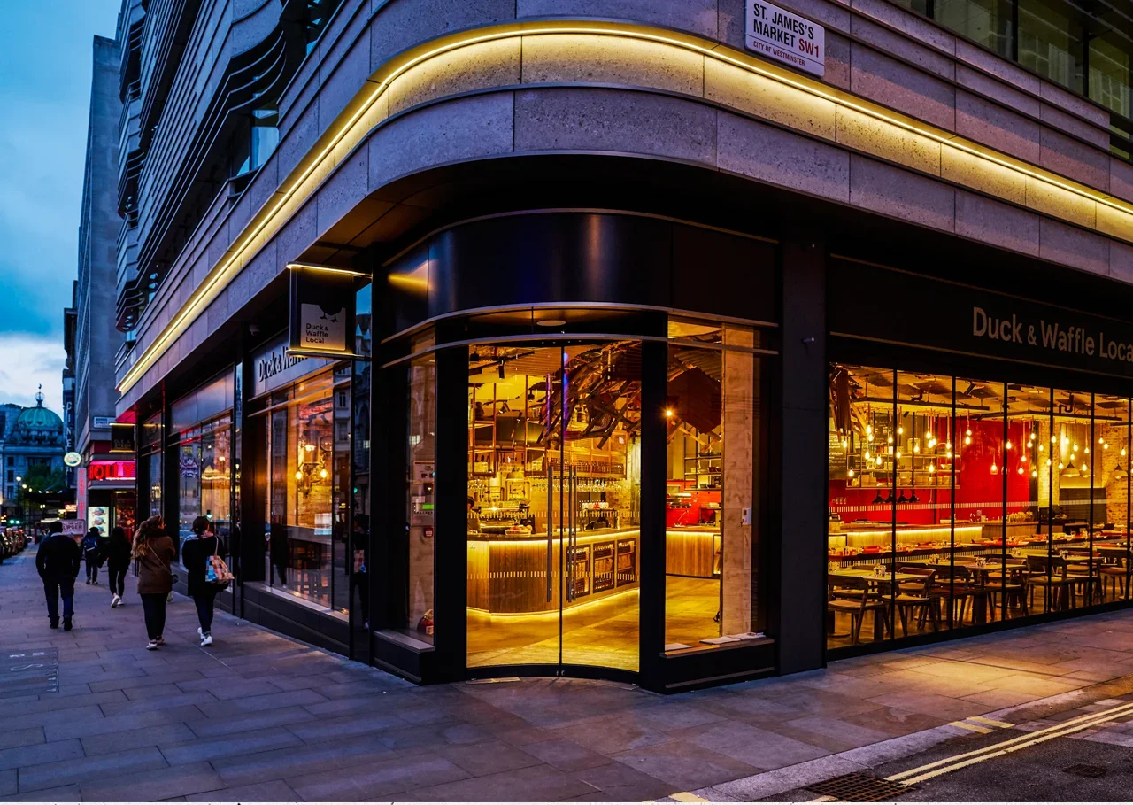 Nighttime street scene showing a modern restaurant with large glass windows and warm interior lighting, located on a city corner with pedestrians walking by.