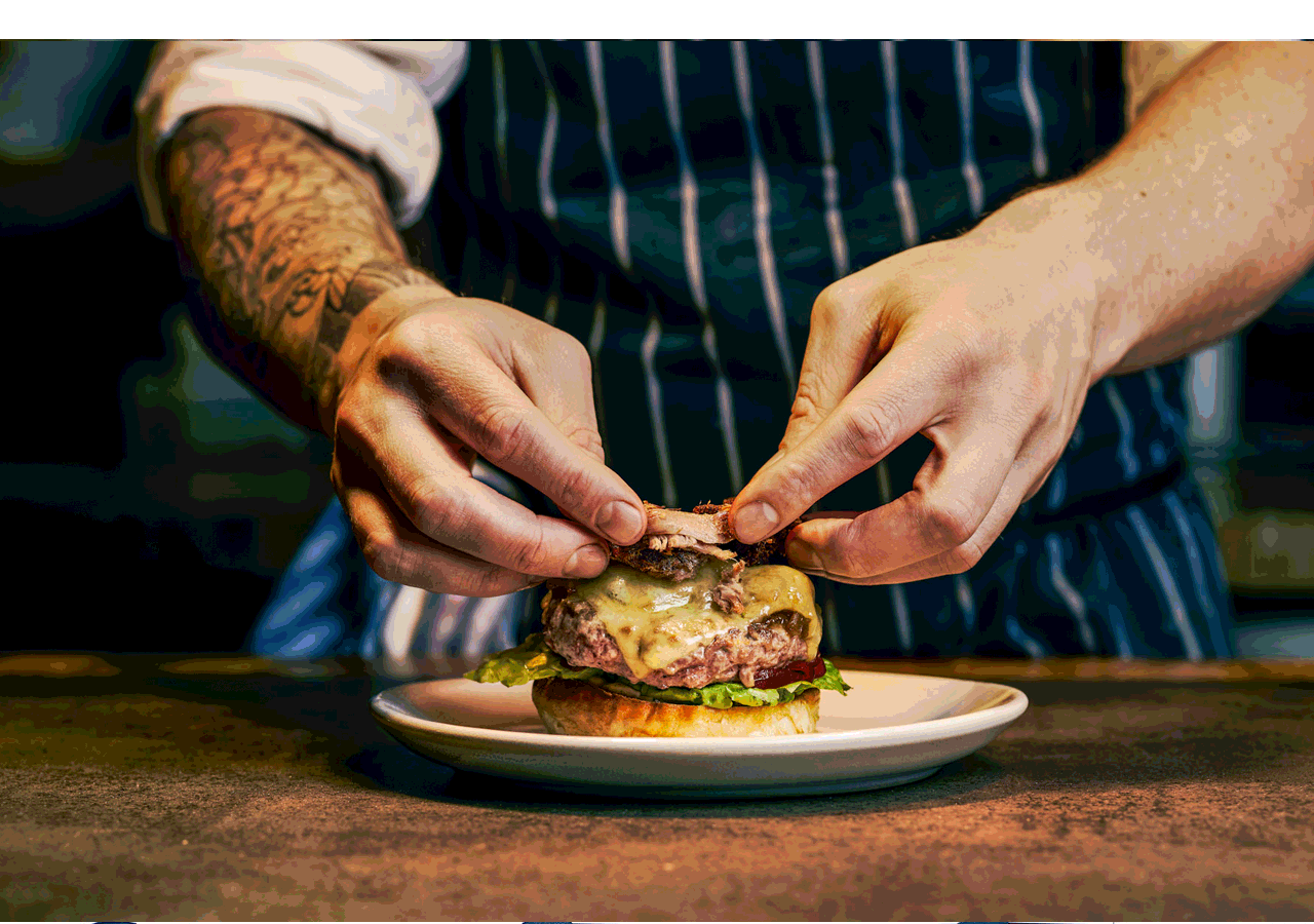 Person assembling a cheeseburger with lettuce, beef patty, melted cheese, and other toppings on a plate.