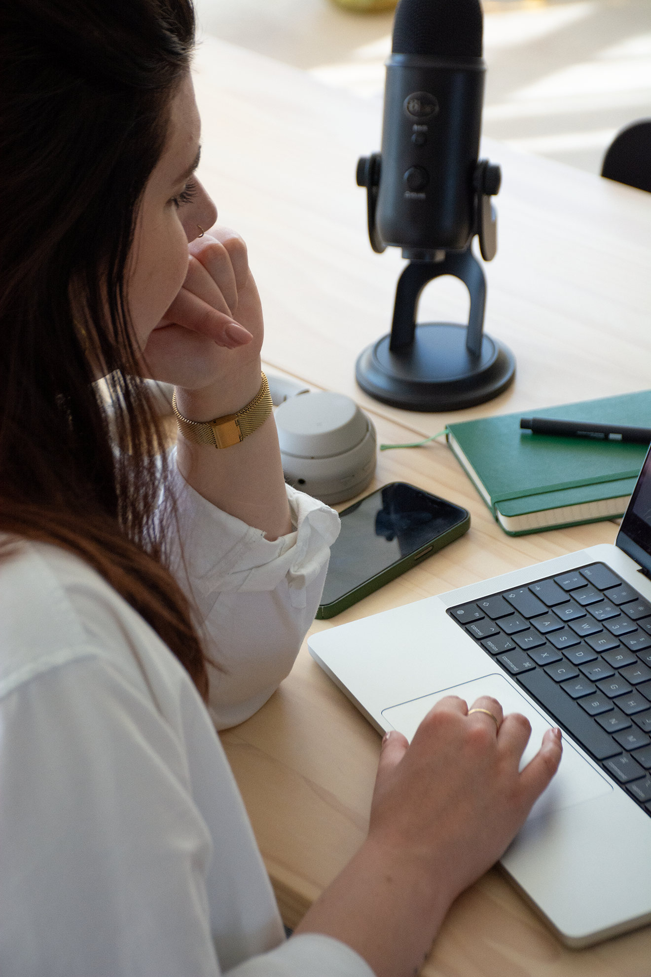 Vrouw die op een laptop typt, omringd door een microfoon, een groene notitieboek, een smartphone, en een pen op een bureau.