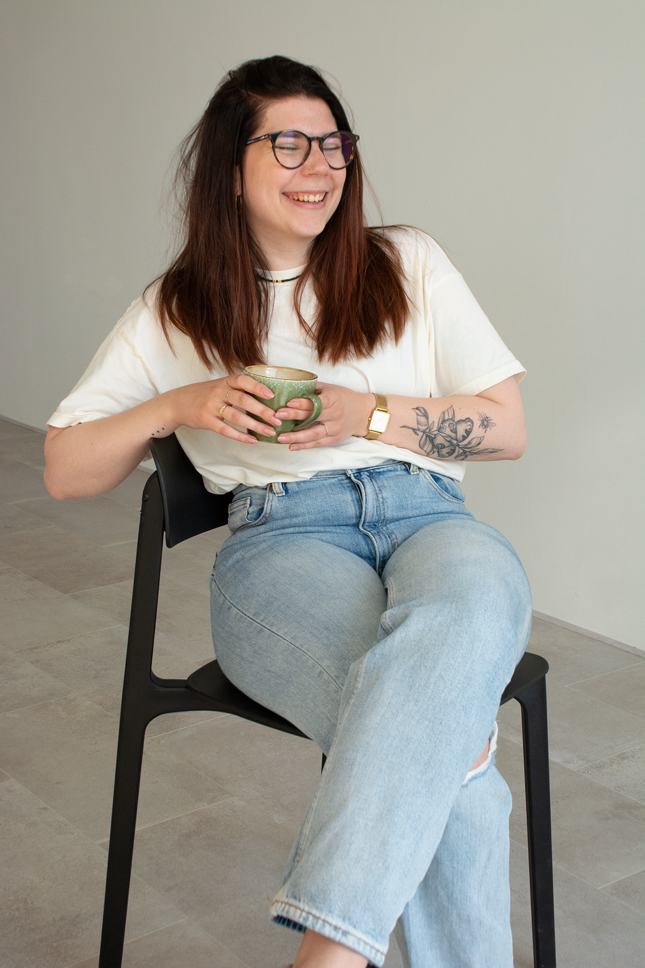 Vrouw met bruin haar en glazen, lachend en zittend op een zwarte stoel, terwijl ze een groene kop vasthoudt, gekleed in een witte T-shirt en spijkerbroek, met een tattoo op haar linkerarm en een horloge om haar linkerpols.