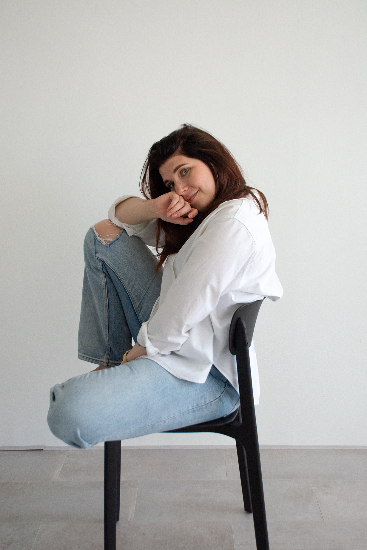 Een jonge vrouw met lang, bruin haar zit op een zwarte stoel, gekleed in een witte blouse en lichtblauwe jeans met een scheur aan de knie. Ze zit zijwaarts met haar rechterbeen omhoog op haar linkerbeen en leunt haar hoofd op haar hand, lachend naar de camera, tegen een witte muur.