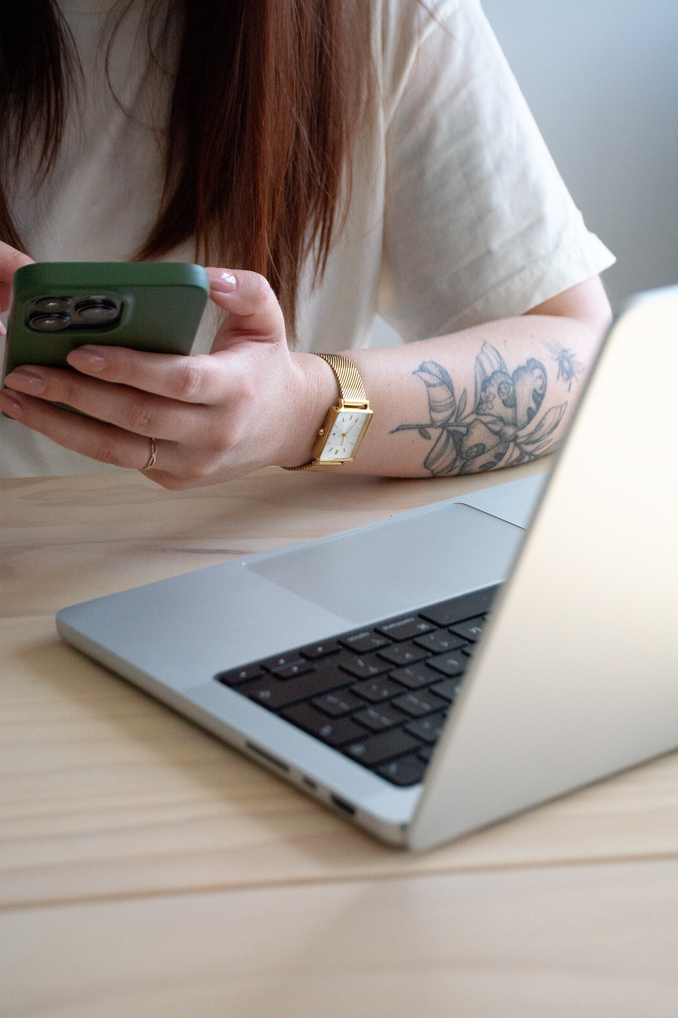 Persoon met rode haren, dragen een wit T-shirt, houdt een groene smartphone en zit voor een laptop op een houten bureau. Ze dragen een gouden horloge en een ring. Op haar linkerarm tatoeages van een bij, blad en insect.