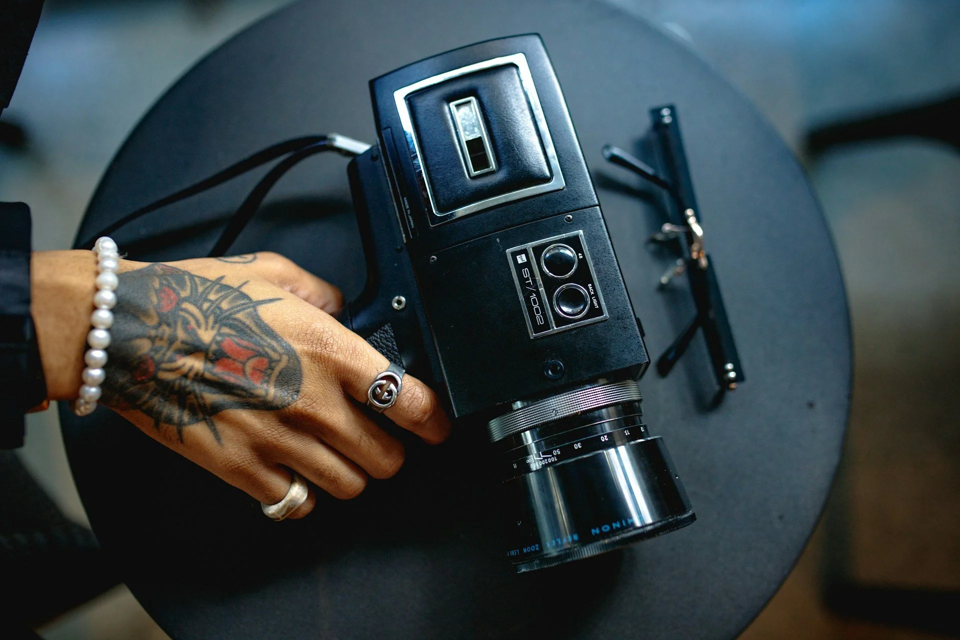 Een hand met tattoos en sieraden houdt een vintage camera vast, rustend op een zwarte stoel.