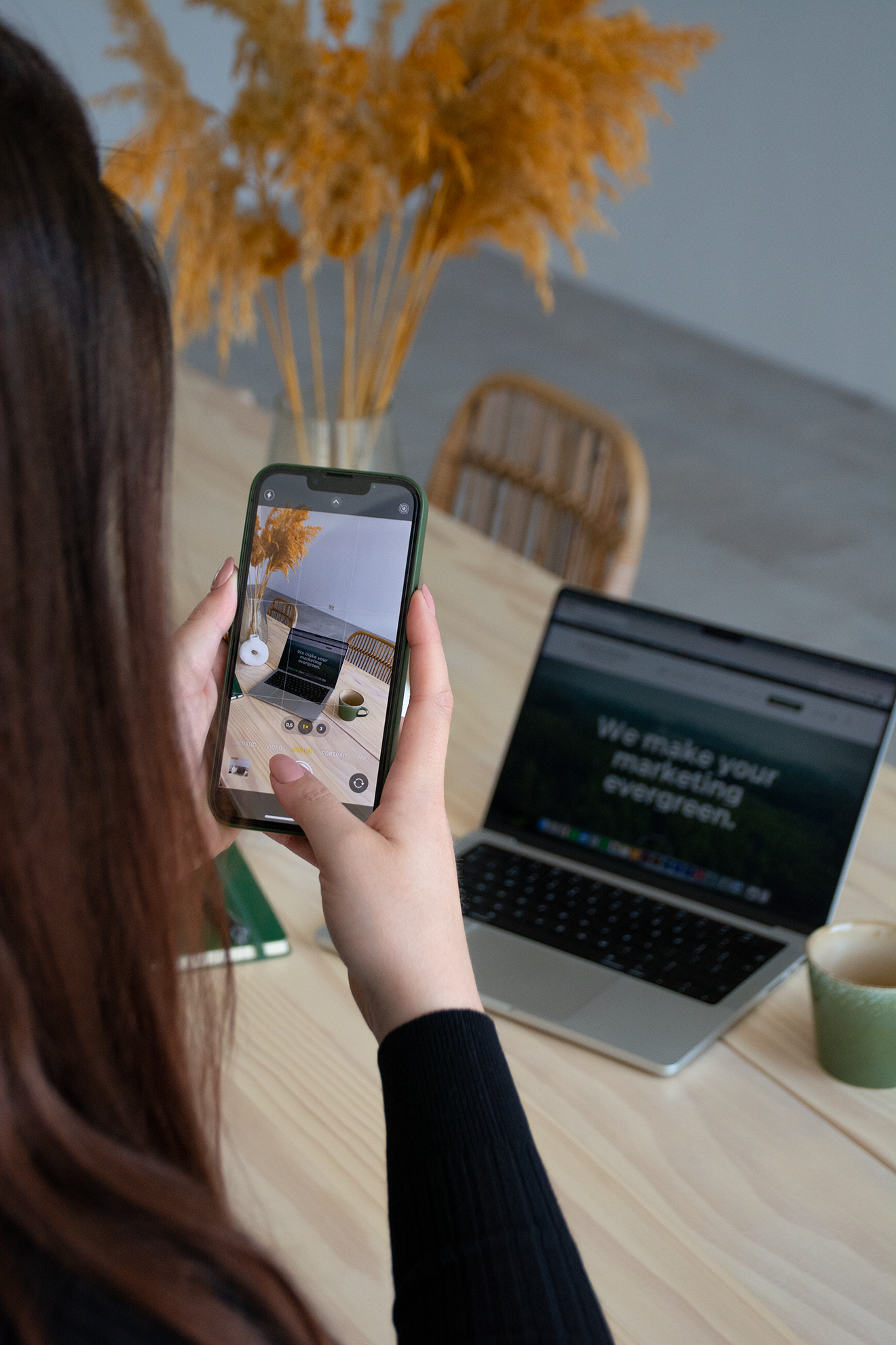 Een persoon maakt een foto van een gedroogde bloem op een houten tafel met een laptop en een kopje koffie.