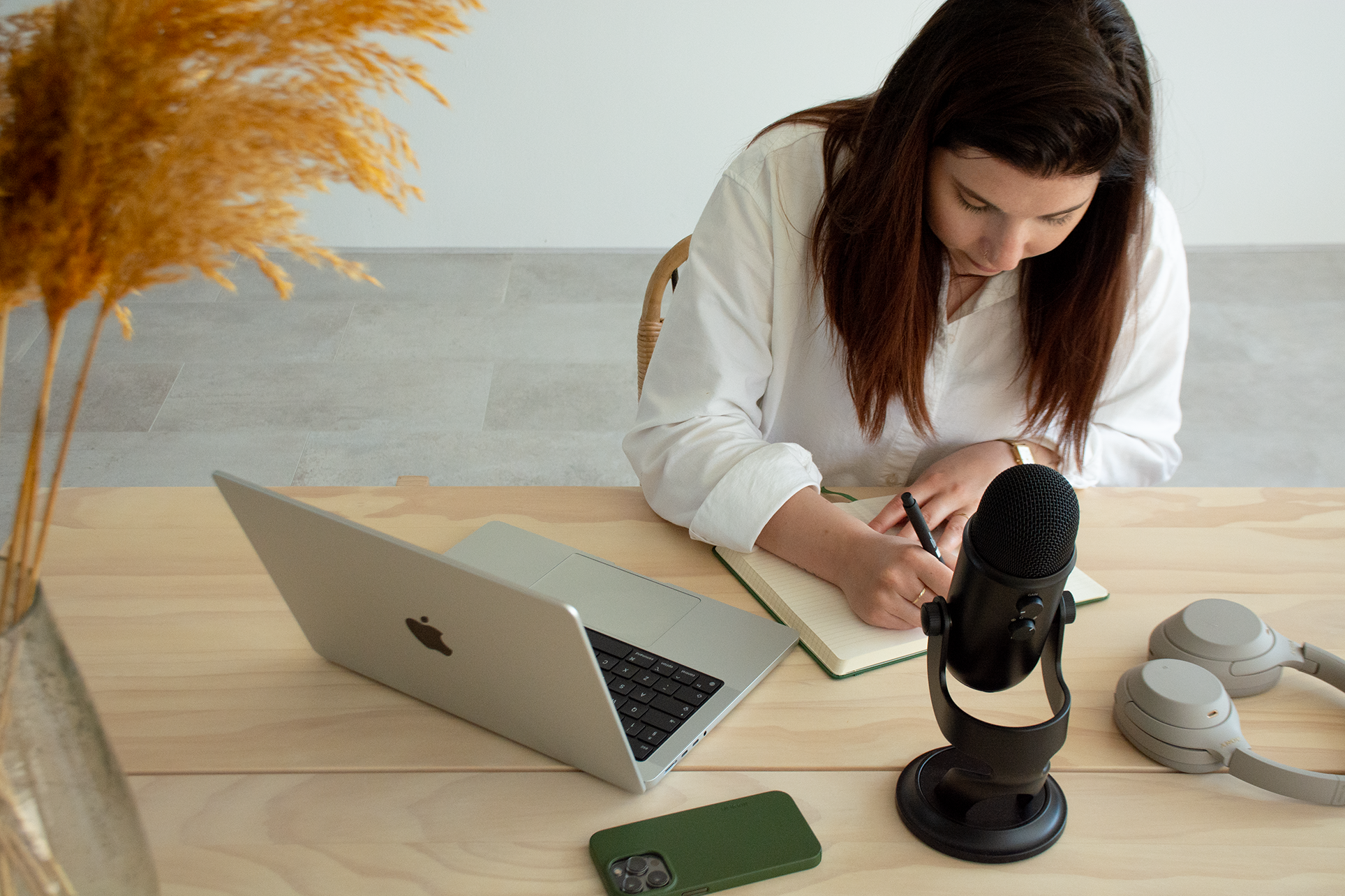 Een vrouw schrijft in een notitieboekje terwijl ze voor een microfoon zit, omringd door een laptop, een iPhone, koptelefoons en andere technische apparatuur op een houten tafel.