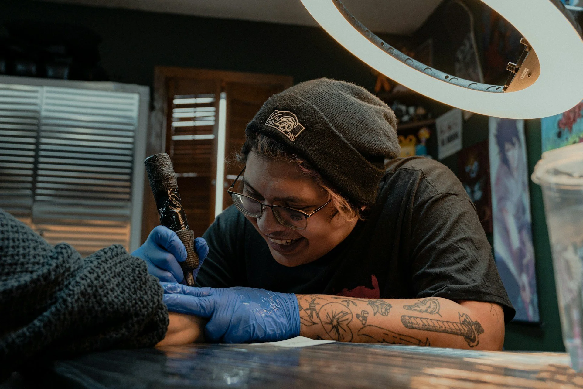 Tattoo-artist die een tattoo zet op de arm van een cliënt, lachend en met een beanie op. Binnen in de studio met verlichte lamp en posters op de muur.
