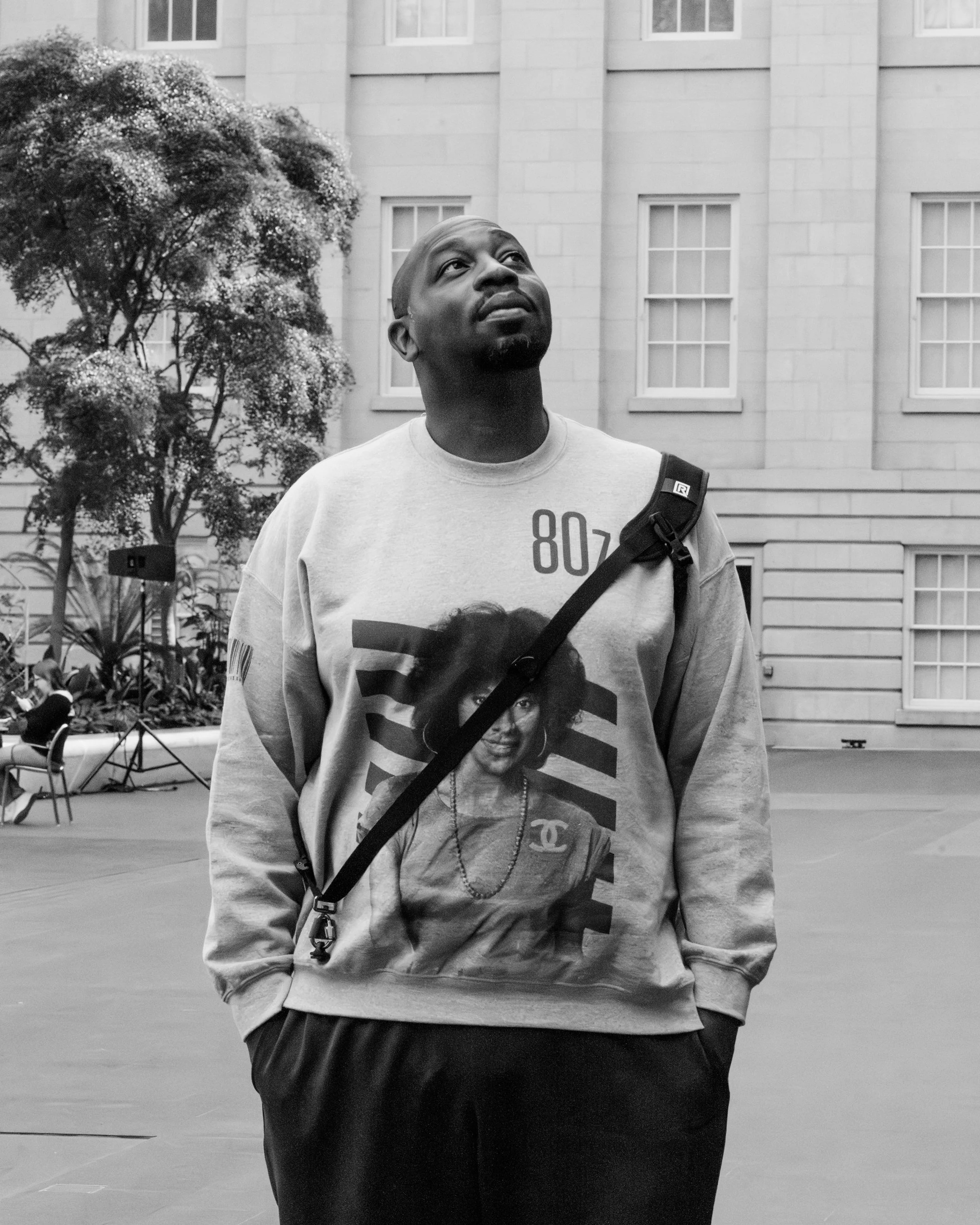 A man standing outdoors, looking up with a thoughtful expression. He is wearing a sweatshirt with a portrait of a woman and the number 80z, and a backpack. There are trees and a building in the background.