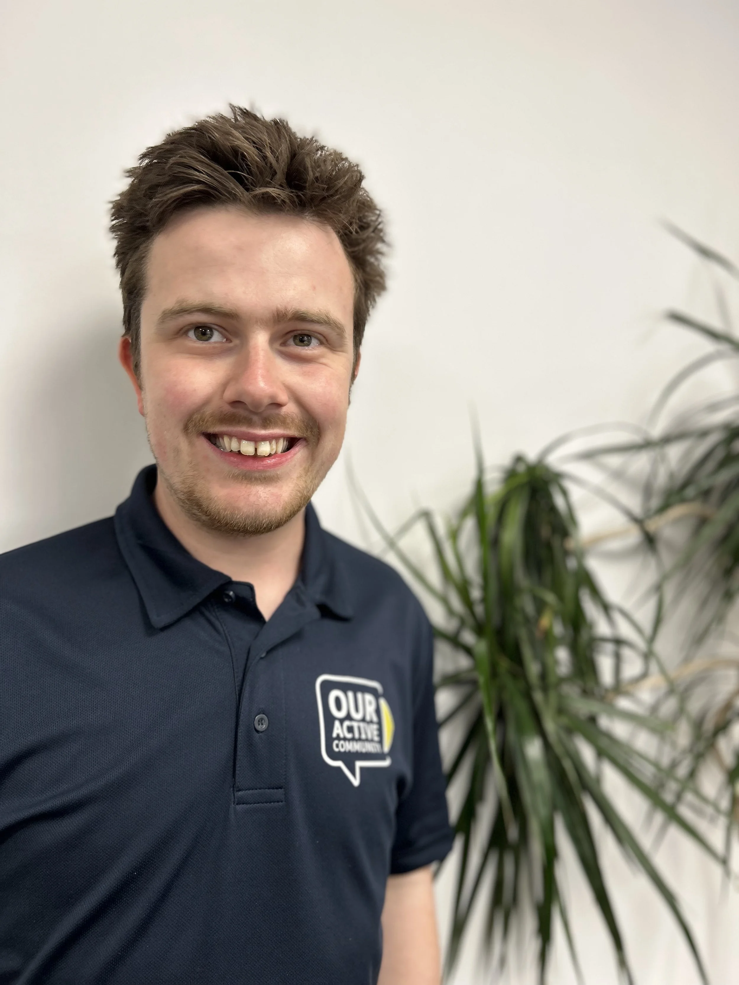 A young man with brown hair and a beard smiling, wearing a dark blue polo shirt with a logo that reads 'OUR ACTIVE COMMUNITY' on the left side, standing in front of a white wall with a large green plant on the right.