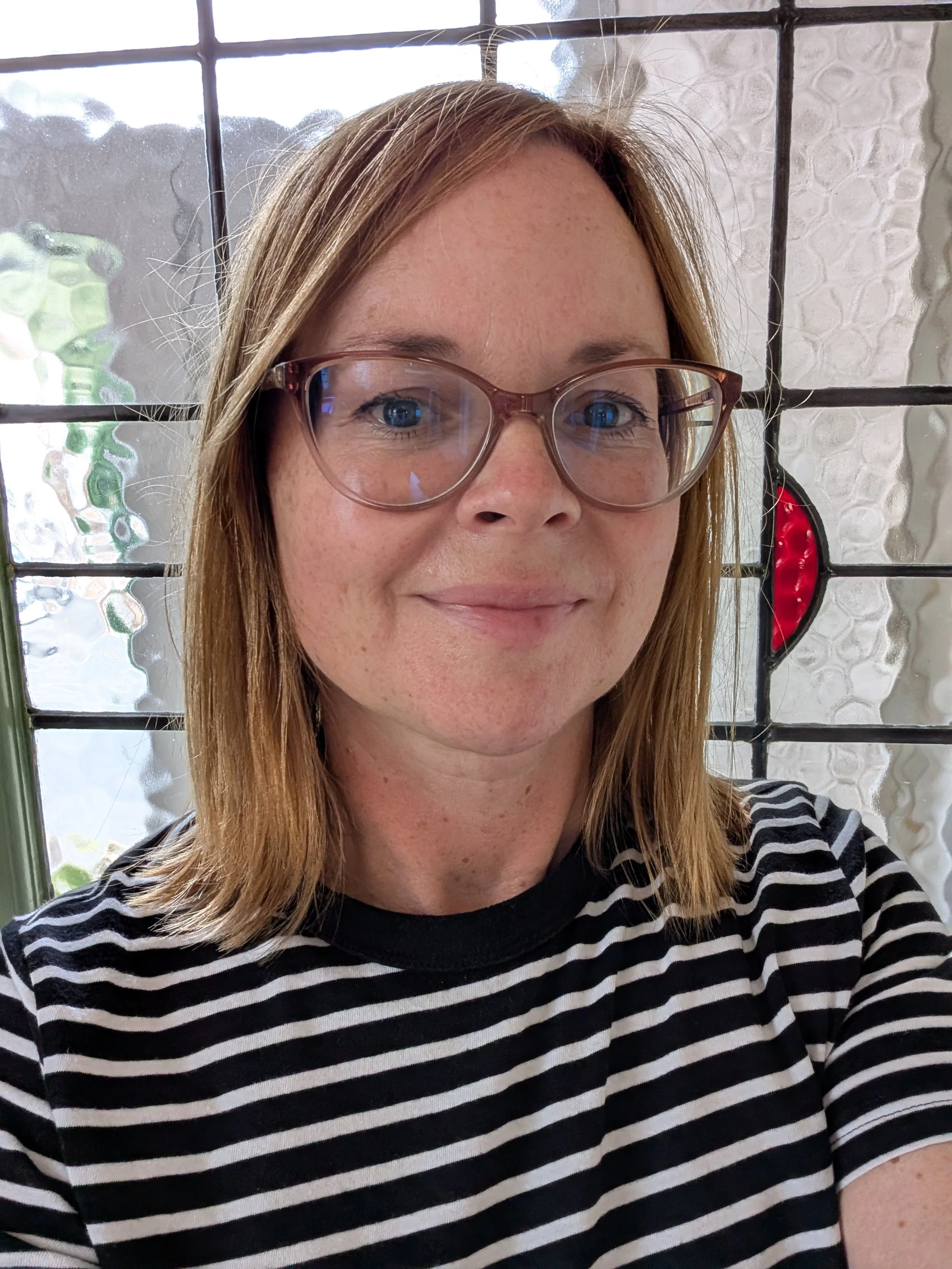 Close-up of a woman with red hair, glasses, wearing a black and white striped shirt, smiling, in front of a textured glass window with a decorative red and black design.