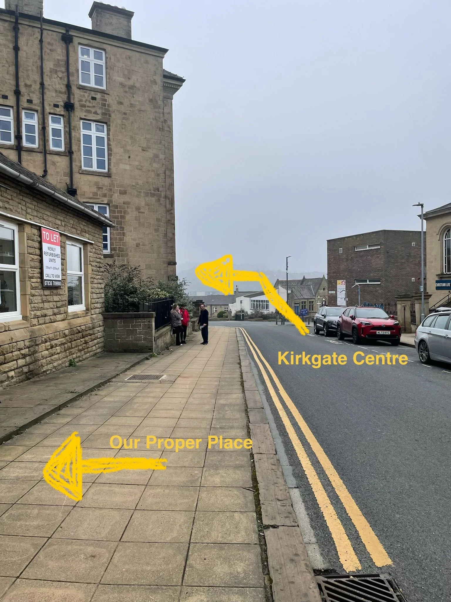 A street view with buildings on the left and parked cars on the right. A yellow arrow points to the left side of the sidewalk labeled 'Our Proper Place,' and another yellow arrow points to a building round the left corner labeled 'Kirkgate Centre.' 
