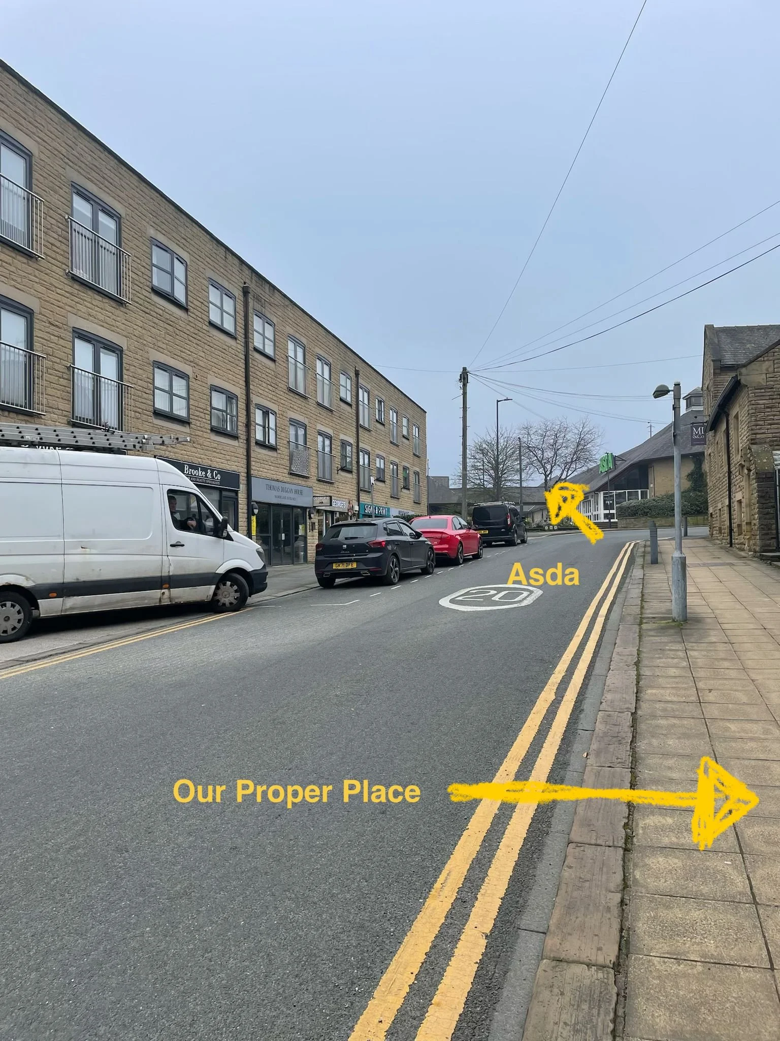A street scene with parked cars along a pavement, a large building with multiple windows. A marked arrow points forwards to Asda, another arrow point to the left with the text Our Proper Place.