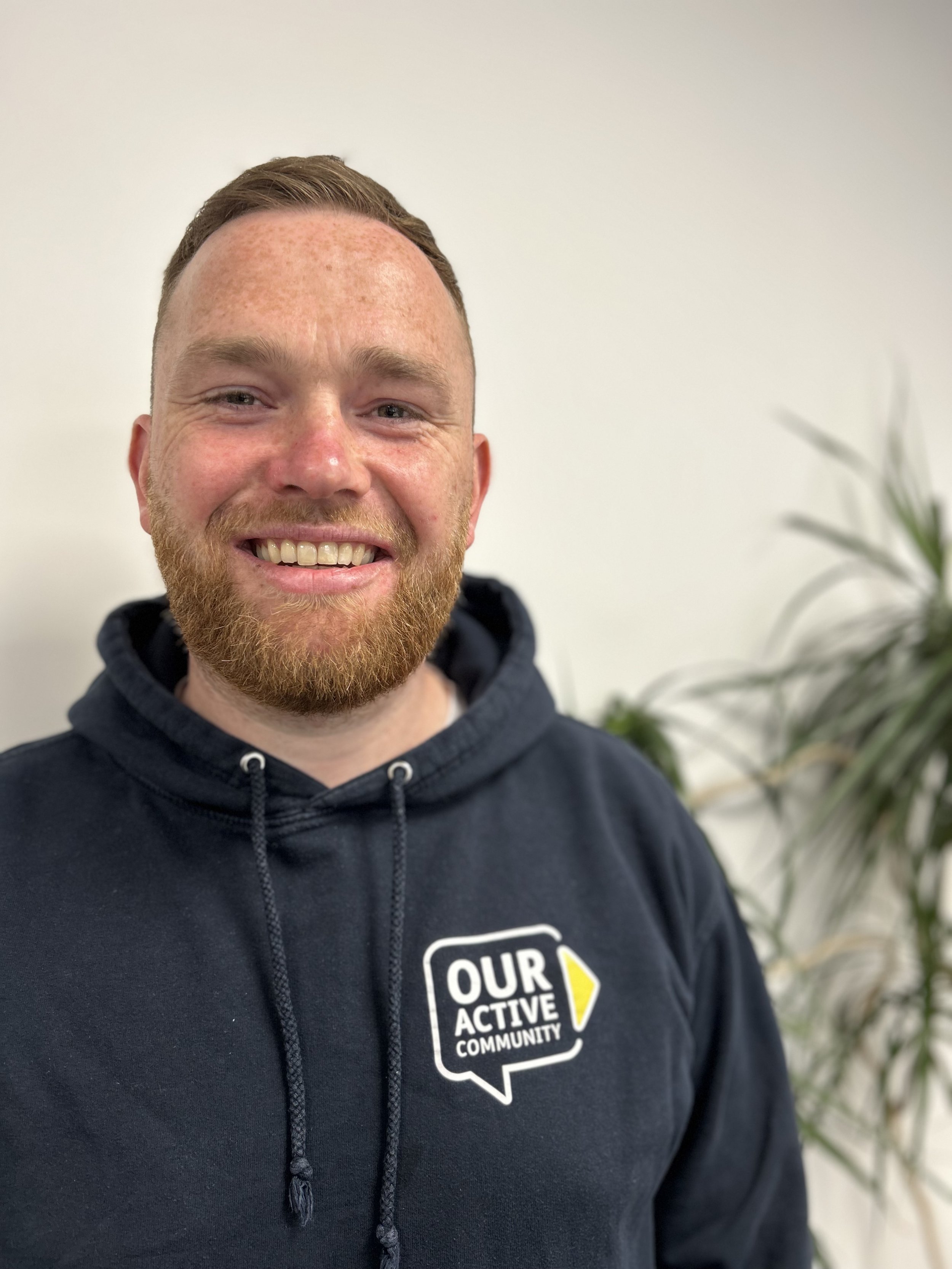 A smiling man with a beard and short hair wearing a black hoodie with a logo that says "Our Active Community" in a speech bubble on the left side of his chest. In the background, there are some green plants and a plain light-colored wall.
