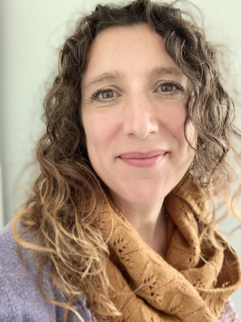 A white, middle aged woman with curly, light brown hair and brown eyes smiling at the camera, wearing a light purple top and a burnt orange, patterned scarf.