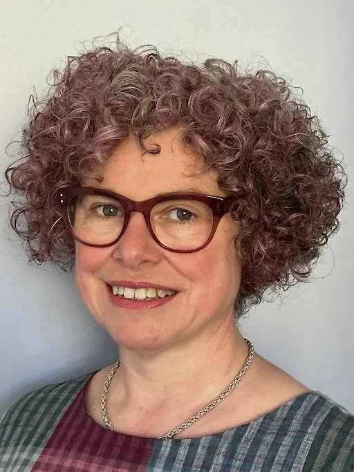 A woman with curly, short gray hair, wearing glasses and a multicolored plaid shirt, smiling in front of a plain light background.