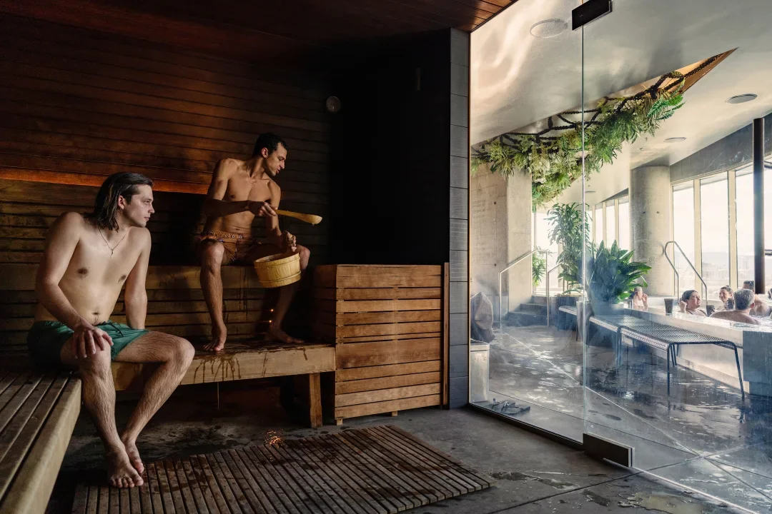 People relaxing in a modern hot tub room with a sauna, where one person is sitting on a bench and another is pouring water over himself with a ladle, while others relax in an adjacent hot tub area visible through glass walls.
