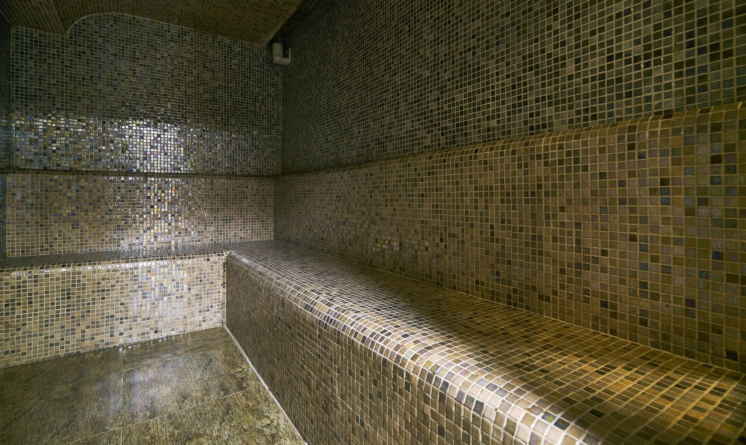 Interior of a steam room with mosaic tiled walls and seating, subdued lighting, and a small ceiling-mounted vent.