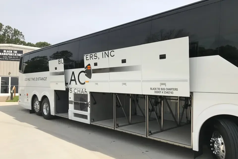 A white and black bus with an open side panel revealing empty storage compartments. The bus has part of a logo and text that reads 'Black Tie Bus Charters' and 'US DOT 2340743'.